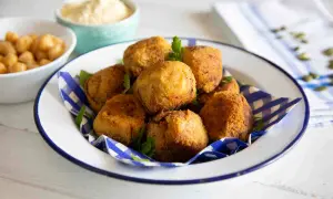Un piatto di polpette di ceci dorate e croccanti, guarnite con prezzemolo fresco. Sullo sfondo, ciotole con ceci e una crema chiara.