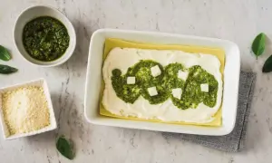 Lasagne al pesto in preparazione: strati di pasta, besciamella, pesto e cubetti di formaggio. Accanto, ciotole con pesto e parmigiano grattugiato.