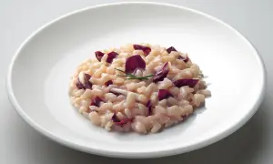 Un cremoso risotto al radicchio, guarnito con foglie viola e un rametto di erba cipollina, servito su un piatto bianco.