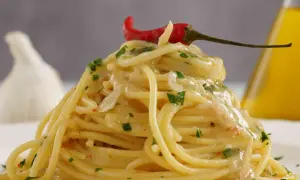 Spaghetti aglio olio e peperoncino cremosi, guarniti con un peperoncino rosso e prezzemolo. Sullo sfondo, aglio e olio d'oliva.
