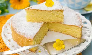 Una fetta di torta paradiso sofficissima, spolverata di zucchero a velo e decorata con un fiore giallo, su un piatto bianco tra fiori arancioni.