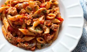 Piatto di pasta alla boscaiola con penne, funghi, salsiccia e pomodoro, guarnito con prezzemolo.