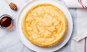 Pila di crepes senza uova dorate su un piatto bianco, con una ciotola di sciroppo d'acero e un dosatore di miele accanto.