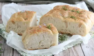 Focaccia senza glutine soffice e dorata, guarnita con rosmarino fresco e piccoli fiori viola, su carta da forno.