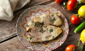 Due fette di pesce spada in friggitrice ad aria, guarnite con prezzemolo, su un piatto decorato. Accanto, pomodori, limoni e zucchine.