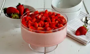 Delizioso mousse di fragole in una coppa di vetro, guarnito con fragole fresche a pezzi. Accanto, una ciotola di fragole e ciotole bianche.
