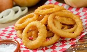Anelli di cipolla dorati e croccanti, preparati con una perfetta pastella per fritti, serviti su carta a scacchi rossi e bianchi, con sale grosso a lato.