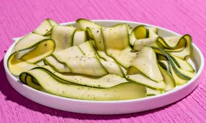 Fettine sottili di zucchine marinate crude, condite con pepe nero, disposte a spirale in un piatto bianco su un tavolo rosa acceso.
