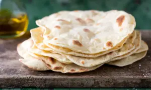 Pila di pane senza lievito, cotto su una tavola di legno grezzo, con un tocco di sale e una bottiglia d'olio sullo sfondo.