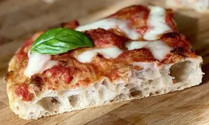 Fetta di pizza in teglia con pomodoro, mozzarella filante e una foglia di basilico fresco. La crosta è alta e alveolata, segno di una lievitazione perfetta.