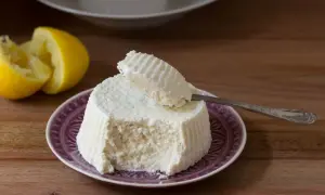 Ricotta fatta in casa su un piatto viola, con limone a fianco. Un cucchiaino preleva una porzione cremosa.