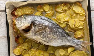 Orata al forno dorata e succulenta, adagiata su un letto di patate affettate e cotte a puntino, profumate di erbe aromatiche.