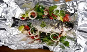 Branzino al cartoccio pronto per la cottura, con pomodorini, cipolla e prezzemolo su foglio d'alluminio.