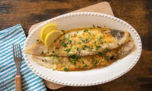 Due sogliole alla mugnaia dorate, guarnite con prezzemolo e spicchi di limone, servite su un piatto bianco.