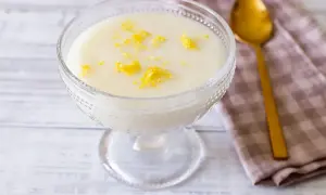 Deliziosa crema di riso in una coppa di vetro, guarnita con scorza di limone. Accanto, un cucchiaio dorato e un tovagliolo a quadretti.
