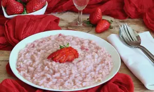 Un cremoso risotto alle fragole, guarnito con una fragola fresca, su un piatto bianco, circondato da fragole rosse e un drappo di seta.