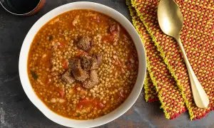 Una ciotola di minestra di lenticchie fumante, guarnita con crostini di pane, accanto a un tovagliolo colorato e un cucchiaio dorato.