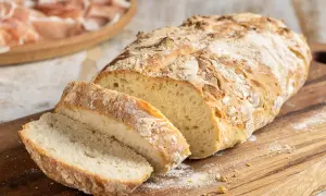 Pane fatto in casa con farina 00, tagliato a fette su un tagliere di legno, con affettati in lontananza.