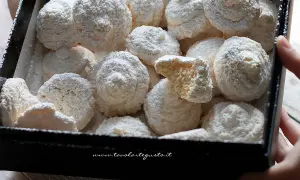 Scatola piena di soffici biscotti al cocco, spolverati di zucchero a velo. Uno è spezzato, rivelando la sua consistenza friabile.