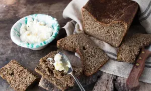 Fetta di pane di segale con burro spalmato, accanto a una ciotola di formaggio fresco e un coltello.
