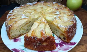 Una fetta di torta di mele, cosparsa di zucchero a velo, su un piatto fiorato. Sullo sfondo, una mela verde.