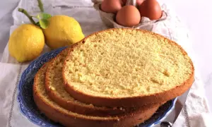 Fette di pan di spagna soffice su un piatto blu, con limoni e uova sullo sfondo, ingredienti per una torta deliziosa.