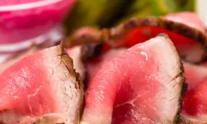 Fette sottili di roast beef ricetta classica, succulente e rosate, con un bordo croccante. Sullo sfondo, un contorno rosa e verde sfocato.