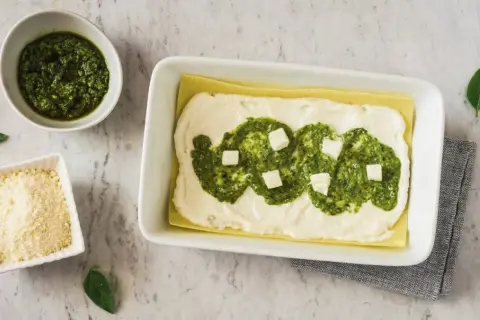 Lasagne al pesto in preparazione: strati di pasta, besciamella, pesto e cubetti di formaggio. Accanto, ciotole con pesto e parmigiano grattugiato.