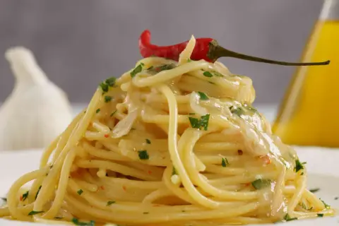 Spaghetti aglio olio e peperoncino cremosi, guarniti con un peperoncino rosso e prezzemolo. Sullo sfondo, aglio e olio d'oliva.