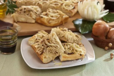 Focaccia con cipolle dorate, fragrante e invitante, servita su un piatto bianco. Sullo sfondo, ingredienti freschi e una bottiglia d'olio.