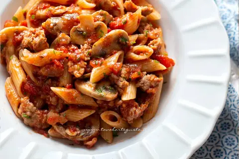 Piatto di pasta alla boscaiola con penne, funghi, salsiccia e pomodoro, guarnito con prezzemolo.