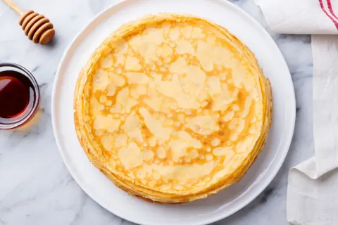 Pila di crepes senza uova dorate su un piatto bianco, con una ciotola di sciroppo d'acero e un dosatore di miele accanto.