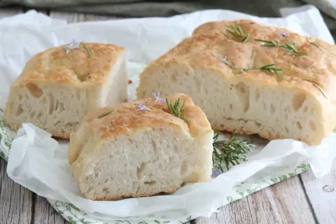 Focaccia senza glutine soffice e dorata, guarnita con rosmarino fresco e piccoli fiori viola, su carta da forno.