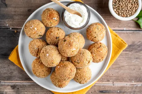 Piatto di polpette di lenticchie dorate e croccanti, servite con una ciotolina di salsa cremosa e un cucchiaino di legno.