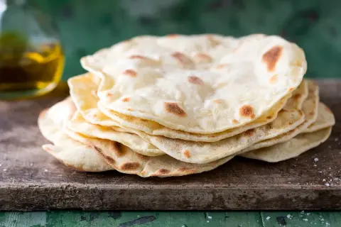 Pila di pane senza lievito, cotto su una tavola di legno grezzo, con un tocco di sale e una bottiglia d'olio sullo sfondo.