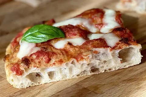 Fetta di pizza in teglia con pomodoro, mozzarella filante e una foglia di basilico fresco. La crosta è alta e alveolata, segno di una lievitazione perfetta.