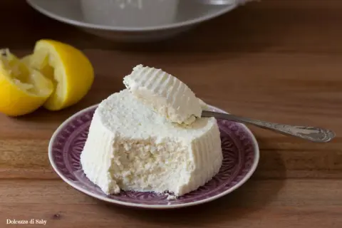Ricotta fatta in casa su un piatto viola, con limone a fianco. Un cucchiaino preleva una porzione cremosa.