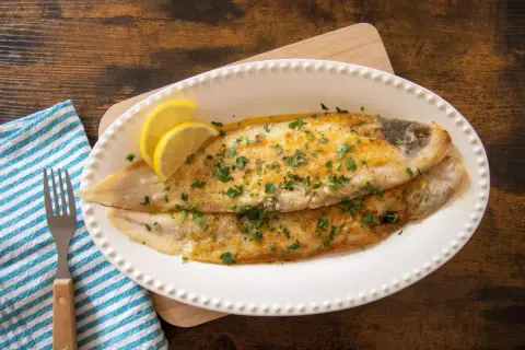 Due sogliole alla mugnaia dorate, guarnite con prezzemolo e spicchi di limone, servite su un piatto bianco.