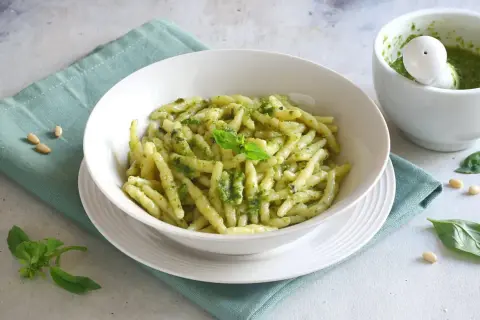 Un piatto invitante di pasta al pesto, condita con basilico fresco e pinoli, accanto a un mortaio con altro pesto.