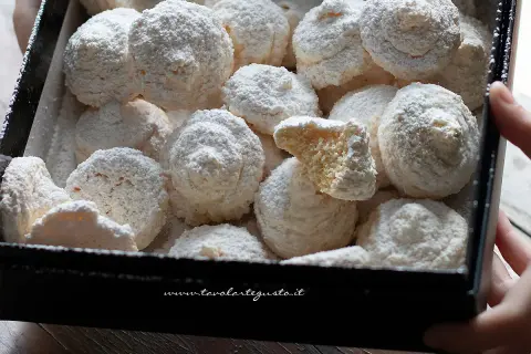 Scatola piena di soffici biscotti al cocco, spolverati di zucchero a velo. Uno è spezzato, rivelando la sua consistenza friabile.