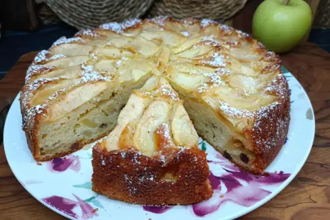 Una fetta di torta di mele, cosparsa di zucchero a velo, su un piatto fiorato. Sullo sfondo, una mela verde.