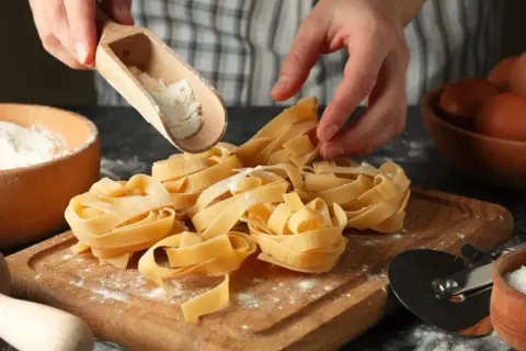 Mani che spolverano farina su nidi di pasta fatta in casa. Ingredienti freschi, uova e sale, pronti per un pasto delizioso.