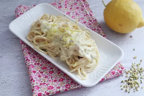 Un piatto di spaghetti al limone cremosi, guarniti con scorza di limone e pistacchi tritati, accanto a un limone intero.