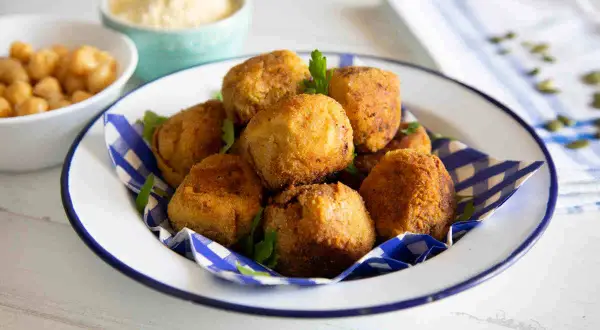 Un piatto di polpette di ceci dorate e croccanti, guarnite con prezzemolo fresco. Sullo sfondo, ciotole con ceci e una crema chiara.