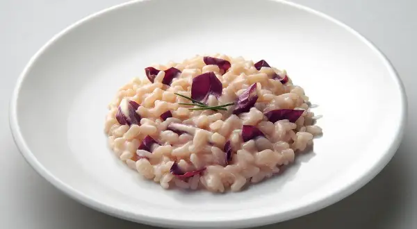 Un cremoso risotto al radicchio, guarnito con foglie viola e un rametto di erba cipollina, servito su un piatto bianco.