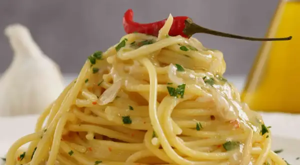 Spaghetti aglio olio e peperoncino cremosi, guarniti con un peperoncino rosso e prezzemolo. Sullo sfondo, aglio e olio d'oliva.
