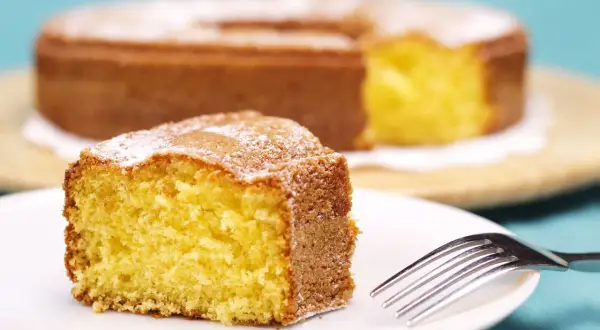 Una fetta di torta 7 vasetti soffice e dorata, spolverata di zucchero a velo, su un piatto bianco con una forchetta.