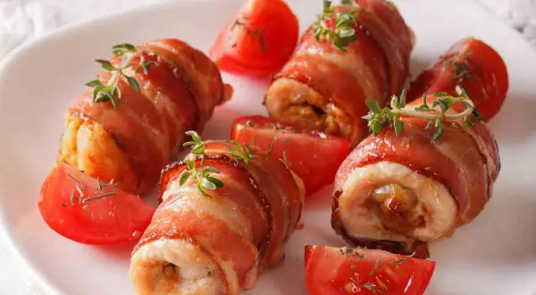 Involtini di pollo avvolti in pancetta croccante, guarniti con timo fresco e serviti con spicchi di pomodoro.