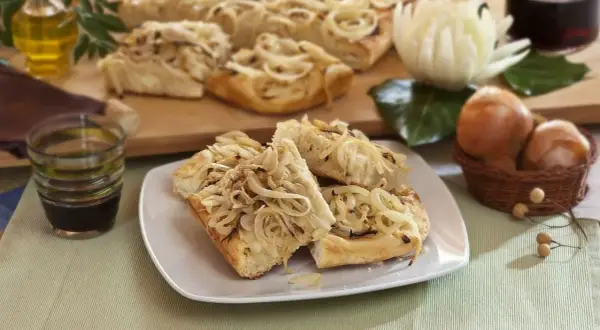 Focaccia con cipolle dorate, fragrante e invitante, servita su un piatto bianco. Sullo sfondo, ingredienti freschi e una bottiglia d'olio.
