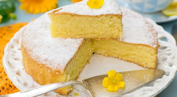 Una fetta di torta paradiso sofficissima, spolverata di zucchero a velo e decorata con un fiore giallo, su un piatto bianco tra fiori arancioni.